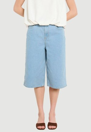 Persona con pantalones cortos de mezclilla azul claro hasta la rodilla, una blusa blanca suelta y tacones marrones abiertos en los dedos, de pie contra un fondo liso.