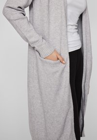 Jasnoszary długi kardigan wykonany z miękkiego materiału, z prążkowanymi mankietami i bocznymi kieszeniami dla dodatkowego detalu i funkcjonalności.