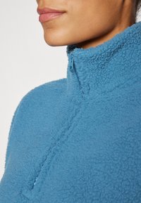 Fleece topp med kvartdragkedja i klarblått, med mjuk textur och hög krage. Dragkedjan matchar plaggets färg.