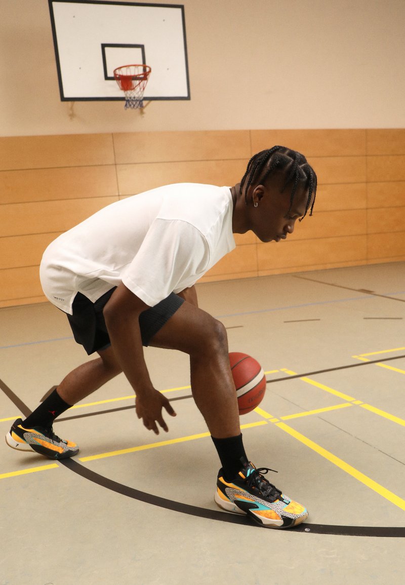 Joueur de basketball dribbler un ballon, portant un t-shirt blanc et un short noir. Ses chaussures présentent un design multicolore avec des motifs et des accents.