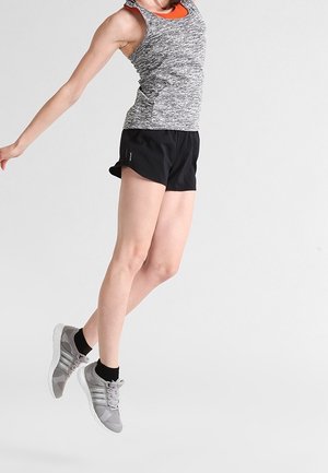 Mujer con camiseta sin mangas gris, pantalones cortos negros y zapatillas grises saltando frente a un fondo blanco liso.