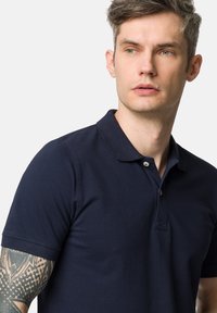 Granatowa koszulka polo wykonana z fakturowanej bawełny. Posiada klasyczny kołnierzyk, dwa guziki i krótkie rękawy. Skóra na przedramieniu widoczna z tatuażami.