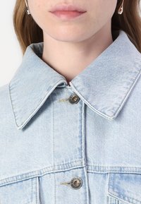 Giacca di jeans azzurro chiaro con un grande colletto, bottoni in metallo argentato e un sottile motivo testurizzato sul tessuto.