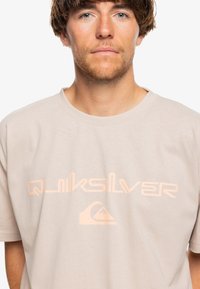 Béžové bavlněné tričko s výrazným světle oranžovým logem "Quiksilver" a grafikou vlny. Klasický kulatý výstřih a krátké rukávy.