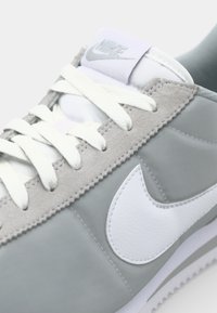 Nike sneakers featuring a gray synthetic upper with suede accents, white logo, and flat white laces. Textured surface, sleek profile.