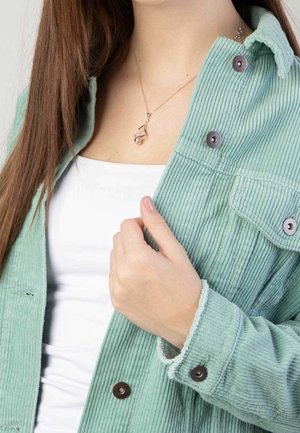 Femme portant une veste en velours côtelé vert pâle sur un haut blanc, tenant le revers de la veste avec la main gauche, et un pendentif en argent visible.