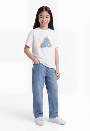 Glunderend meisje met lang zwart haar draagt een wit T-shirt met oranje en blauw driehoeklogo, losse blauwe jeans en witte sneakers.