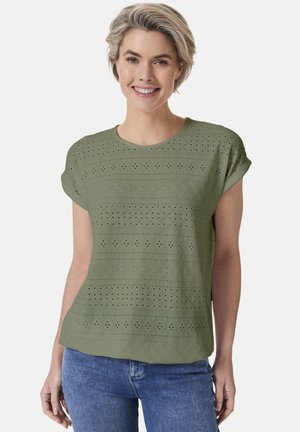 Grünes Kurzarmshirt mit rundem Ausschnitt, das über strukturierte Muster und Lochdetails verfügt. Kombiniert mit blauen Jeans.