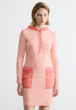 Žena nosi dlhé svetloružové šaty s kapucňou s koralovým gombíkovým prúžkom, kapucňou a dvoma veľkými koralovými prednými vreckami, stojaca na bielom pozadí.