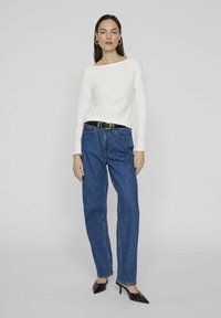 Maglietta bianca a maniche lunghe con texture, jeans blu a gamba dritta, cintura nera con fibbia dorata, tacchi a punta neri. Sfondo semplice.