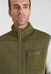 Olivgrüner Außenschutzweste mit einer Mischung aus strukturiertem Fleece und glattem Stoff. Verfügt über eine mit Reißverschluss versehenen Brusttasche und einen hohen Kragen.
