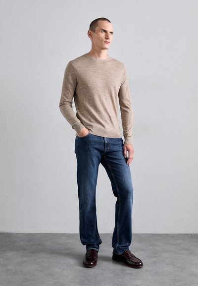 Pull en tricot marron clair, jeans bleus, chaussures marron foncé. Le pull a un col rond et des poignets ajustés. Les jeans sont à coupe droite.