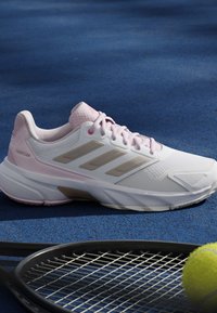 Hvid og lyserød tennissko på blå bane ved siden af en sort tennisketcher og en gul tennisbold.