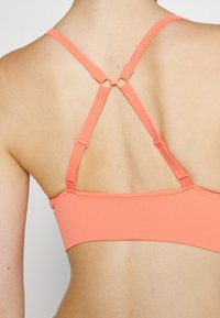 Soutien desportivo de coral ajustável com um design de malha texturizada. Apresenta um padrão de alças cruzadas e um fecho em metal nas costas para suporte.