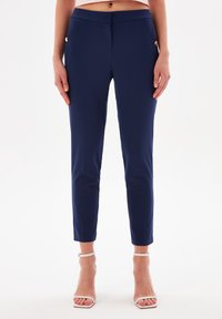 Pantaloni neri elasticizzati con taglio slim, dotati di tasche laterali e dettagli in metallo dorato, texture liscia e lunghezza alla caviglia.