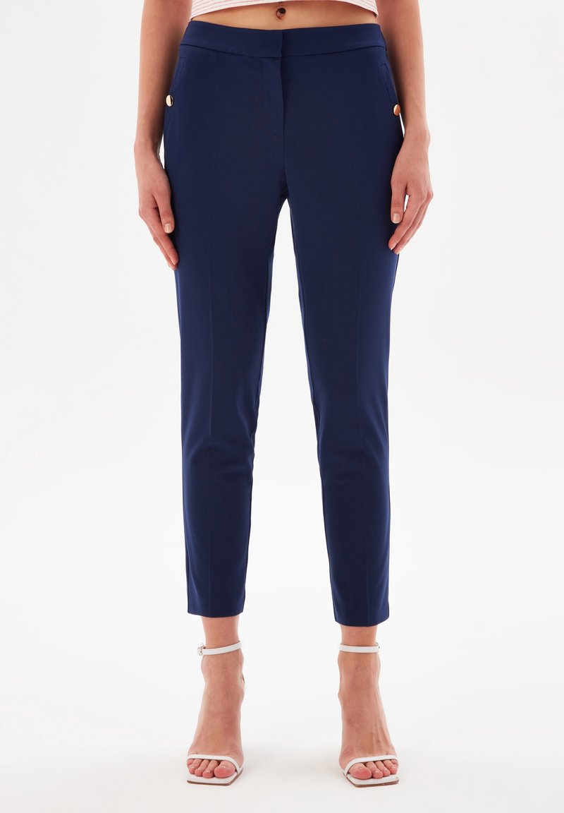 Pantaloni neri elasticizzati con taglio slim, dotati di tasche laterali e dettagli in metallo dorato, texture liscia e lunghezza alla caviglia.