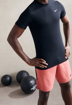 Athlète portant un t-shirt noir Nike et un short corail, debout avec les mains sur les hanches près de trois ballons médicinaux noirs posés au sol.