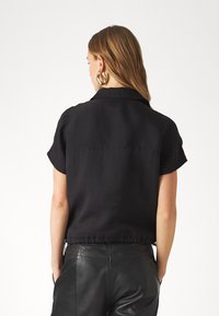 Blouse noire à manches courtes avec col, présentant une texture subtile et un design court, associée à un pantalon en cuir noir.