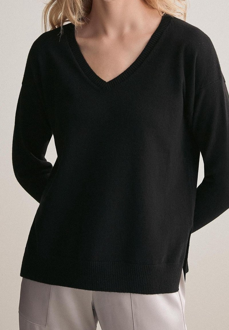 Femme portant un pull noir à col en V manches longues et un pantalon gris clair, debout les mains derrière le dos.