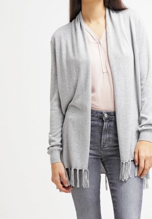 Femme portant un cardigan gris clair ouvert sur le devant avec un ourlet à franges sur une blouse rose pâle et un jean gris, debout, bras détendus.