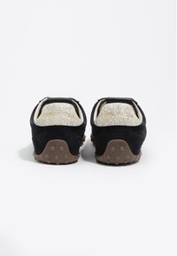 Zapatos de ante negros con un tacón texturizado en color crema, que presentan una suela de goma con agarraderas circulares y sutiles letras doradas en la parte trasera.