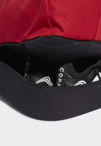 Bolsa de tela roja y negra con cierre con cremallera, que contiene zapatos de fútbol negros con acentos blancos y correas de marca visibles en el interior.