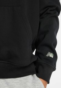 Sudadera negra hecha de tela suave con un bolsillo tipo canguro. Presenta puños y dobladillo acanalados, con un logo reflectante de New Balance en la manga.
