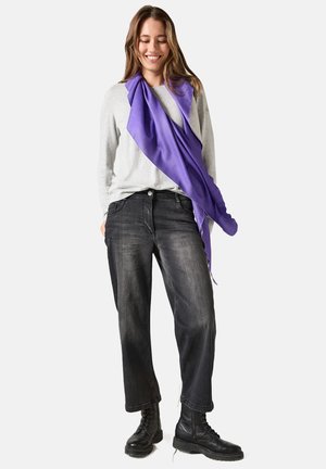Camicia grigia a maniche lunghe, jeans neri larghi e stivali da combattimento neri. Una sciarpa viola si avvolge attorno al collo, aggiungendo un tocco di colore.