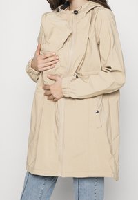 Krémová těhotenská bunda s předním panelem, kterou drží osoba, v kombinaci se světlomodrými džíny.