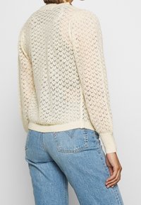 Kremowy sweter z dzianiny z wzorem koronkowym, ściągaczami na mankietach i u dołu oraz luźnym krojem. Połączony z jasnoniebieskimi dżinsami.