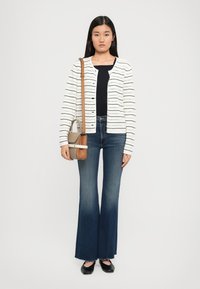 Woman wearing a white striped cardigan, black top, dark flared jeans, black flats, and carrying a beige and brown crossbody bag.