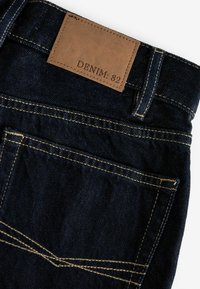Poche arrière de jean en denim bleu foncé avec une étiquette en cuir beige cousue au-dessus affichant le texte "DENIM : 82" et une couture orange visible.