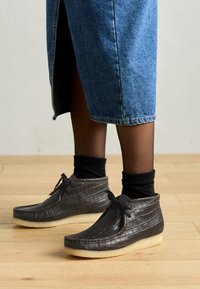 Zwarte schoenen met krokodillenstructuur, voorzien van een ronde neus en een zachte beige zool, vastgemaakt met zwarte veters, gecombineerd met denim en zwarte sokken.