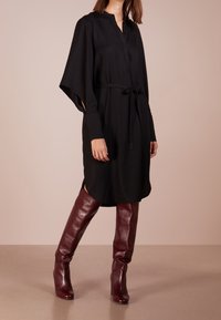 Vestido túnica negro con cuello, mangas largas y cinturón atado en la cintura; combinado con botas de tacón hasta la rodilla de charol burdeos.