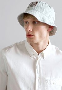 Light gray bucket hat with a stitched logo patch, cotton material, textured surface, and wide brim. Worn with a white button-up shirt.