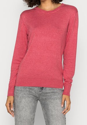 Kobieta w prostym różowym swetrze z długim rękawem i jasnoszarych jeansach w stylu acid wash, stojąca z rozluźnionymi rękoma wzdłuż boków.