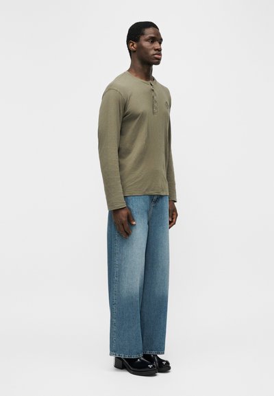 Άνδρας που στέκεται, φοράει ελιάς μακρυμάνικο πουκάμισο henley, φαρδιά ξεθωριασμένα μπλε τζιν και μαύρα μποτάκια με τακούνι, σε απλό λευκό φόντο.