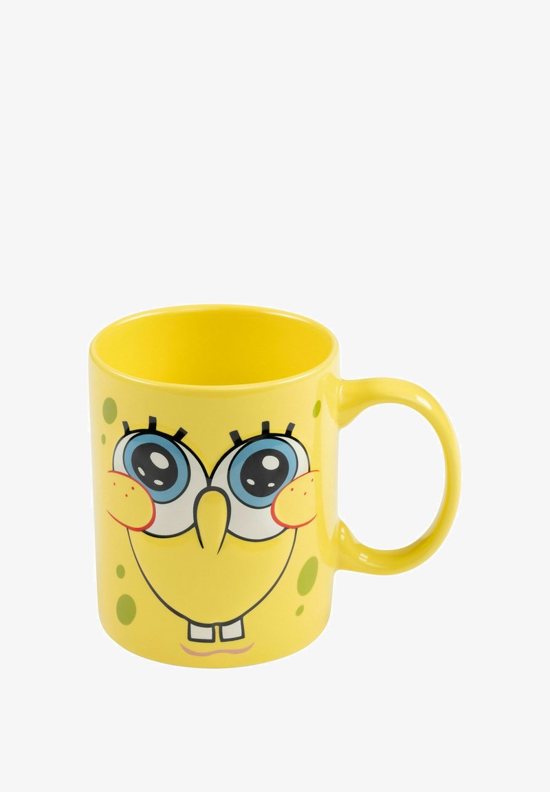 Tazza in ceramica gialla con un volto da cartone animato, grandi occhi, bocca sorridente e macchie di un verde chiaro. Forma arrotondata con un manico laterale.
