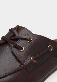 Κοντινή λήψη σκούρου καφέ δερμάτινου παπουτσιού με κορδόνια από το ίδιο δέρμα, περασμένα μέσα από χρυσά μεταλλικά λουράκια.