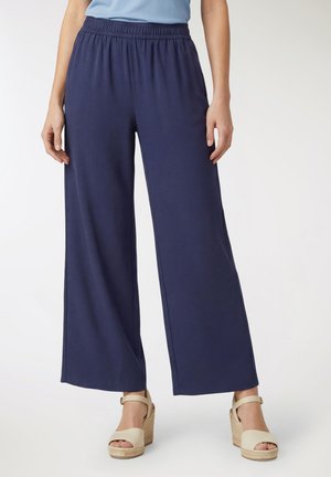Donna che indossa pantaloni larghi blu navy con vita elastica e sandali beige con zeppa, in piedi davanti a uno sfondo uniforme.