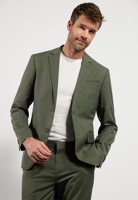 Oliven grøn jakkesæt med matchende bukser, med slim fit, to knapper og en brystlomme. Båret over en hvid T-shirt.