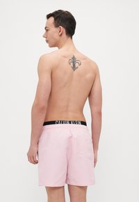 Rosa Badeshorts mit einem schwarzen Calvin Klein Bund, strukturiertem Stoff und einer Gesäßtasche. Ein Tattoo in Form einer Lilie ist sichtbar.