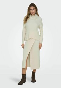 Suéter de punto acanalado color beige claro con cuello alto y falda midi a juego con una abertura lateral. Botines marrones completan el atuendo.