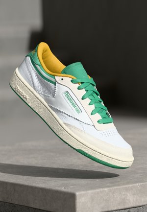 Fehér bőr sportcipő zöld kiegészítőkkel és sárga belsővel, textúrázott talppal és perforált díszítéssel a lábujjaknál. Reebok logó látható.