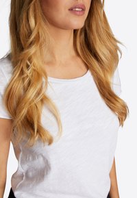 Wit T-shirt met korte mouwen en een ronde halslijn, gemaakt van licht gestructureerde stof met een gladde afwerking. Haar in zachte, losse golven.