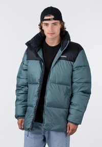 Veste rembourrée à motif bleu-vert avec un panneau supérieur noir. Fermeture éclair sur le devant, poignets élastiques et logo sur la poitrine gauche. Coupe décontractée.
