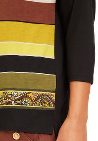 Haut noir avec un ourlet à motifs featuring des rayures marron, jaune et verte. Tissu en mélange de coton et manches 3/4. Fentes latérales visibles.