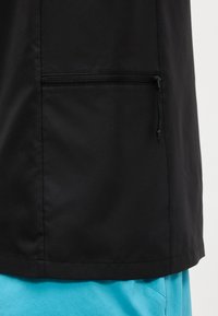 Jachetă neagră cu buzunar orizontal cu fermoar purtată peste pantaloni albastru deschis, arătând trunchiul și zona superioară a piciorului.