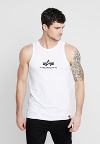 Hvit tank topp med en logo med teksten "ALPHA INDUSTRIES" i svart. Modellen har en tatovering på høyre arm og har på seg svarte bukser.