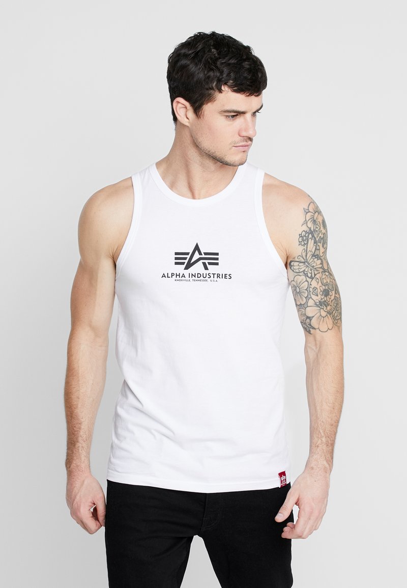 Hvit tank topp med en logo med teksten "ALPHA INDUSTRIES" i svart. Modellen har en tatovering på høyre arm og har på seg svarte bukser.